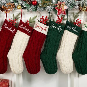May include: Six personalized Christmas stockings in red, white, and green, hanging from a decorated mantel. Each stocking is cable-knit with a white cuff and a name embroidered in white or green. Festive holiday decor surrounds the stockings.