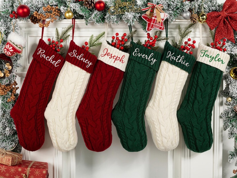 May include: Six personalized Christmas stockings in red, white, and green, hanging from a decorated mantel. Each stocking is cable-knit with a white cuff and a name embroidered in white or green. Festive holiday decor surrounds the stockings.