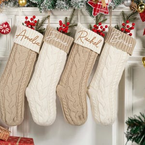 May include: Four cable-knit Christmas stockings in beige and white hang from a decorated doorway. Each stocking has a name embroidered on the cuff: Amelia, Jamie, Rachel, and Bruce. Red berries and greenery adorn the tops.