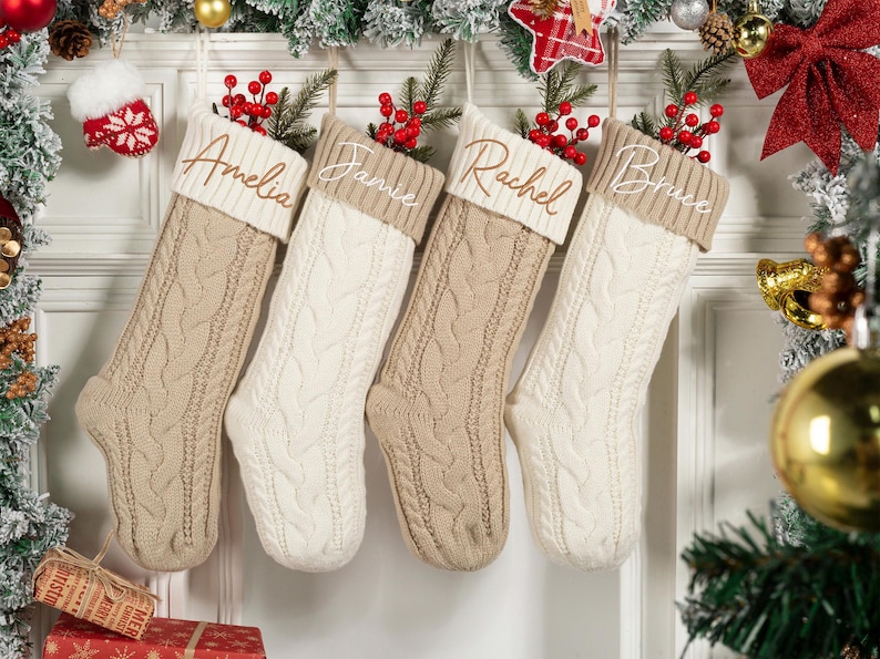 May include: Four cable-knit Christmas stockings in beige and white hang from a decorated doorway. Each stocking has a name embroidered on the cuff: Amelia, Jamie, Rachel, and Bruce. Red berries and greenery adorn the tops.