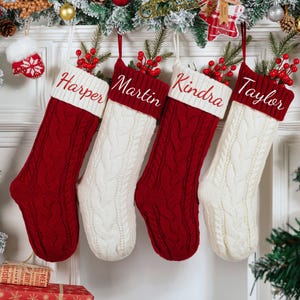 May include: Four personalized Christmas stockings hang, each with a name embroidered on the cuff: Harper, Martin, Kindra, and Taylor. The stockings alternate between red and white cable knit, adorned with berries and greenery.