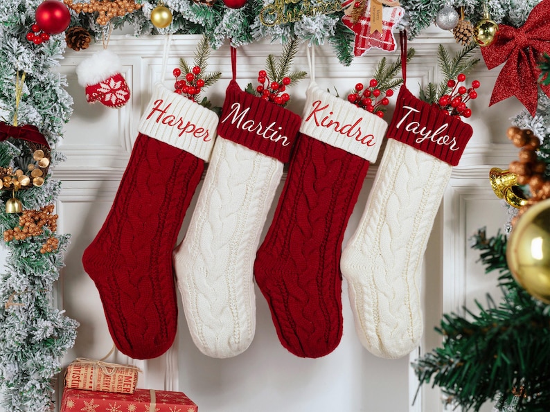 May include: Four personalized Christmas stockings hang, each with a name embroidered on the cuff: Harper, Martin, Kindra, and Taylor. The stockings alternate between red and white cable knit, adorned with berries and greenery.