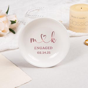Pode incluir: Um prato redondo de cer&acirc;mica branca com letras em ouro rosa que dizem m cora&ccedil;&atilde;o k, ENGAGED e a data 14.02.21. O prato tem um acabamento liso e brilhante com um design circular simples para colocar pequenos objetos.