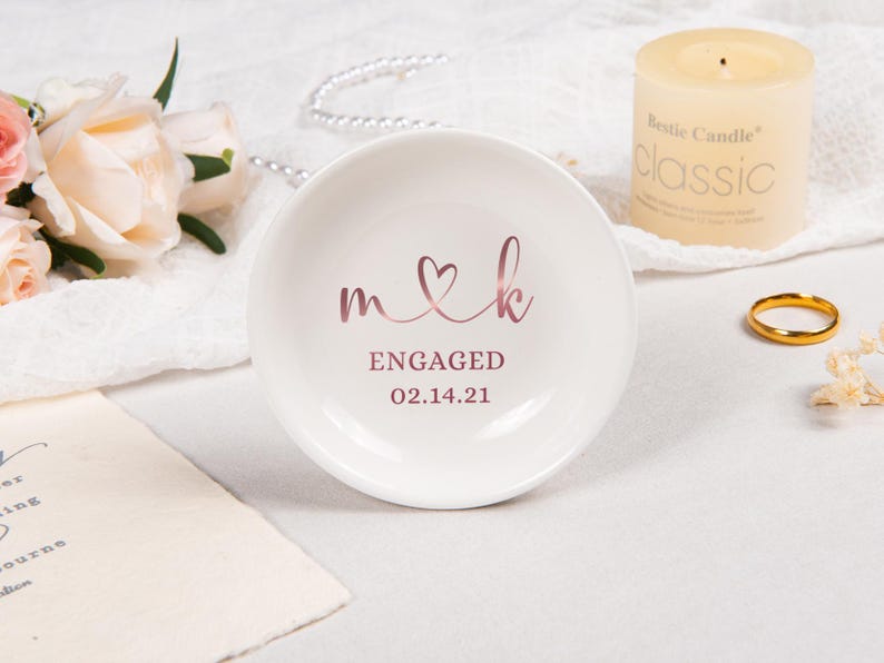 Pode incluir: Um prato redondo de cer&acirc;mica branca com letras em ouro rosa que dizem m cora&ccedil;&atilde;o k, ENGAGED e a data 14.02.21. O prato tem um acabamento liso e brilhante com um design circular simples para colocar pequenos objetos.