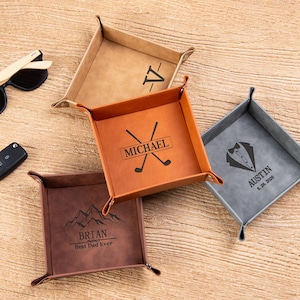 May include: Four square valet trays in tan, orange, dark brown, and grey leather. The tan tray features a V monogram. The orange tray shows the name MICHAEL with golf clubs. The grey tray displays a tuxedo graphic, AUSTIN, and 6. 20. 2026. The dark brown tray shows mountains, BRIAN, and Best Dad Ever.
