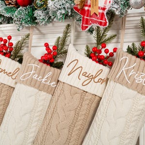 May include: Four cable-knit Christmas stockings in cream and tan, each with a name embroidered on the cuff: Ameli, Jackie, Naylor, and Kristin. The stockings are decorated with red berries and greenery.