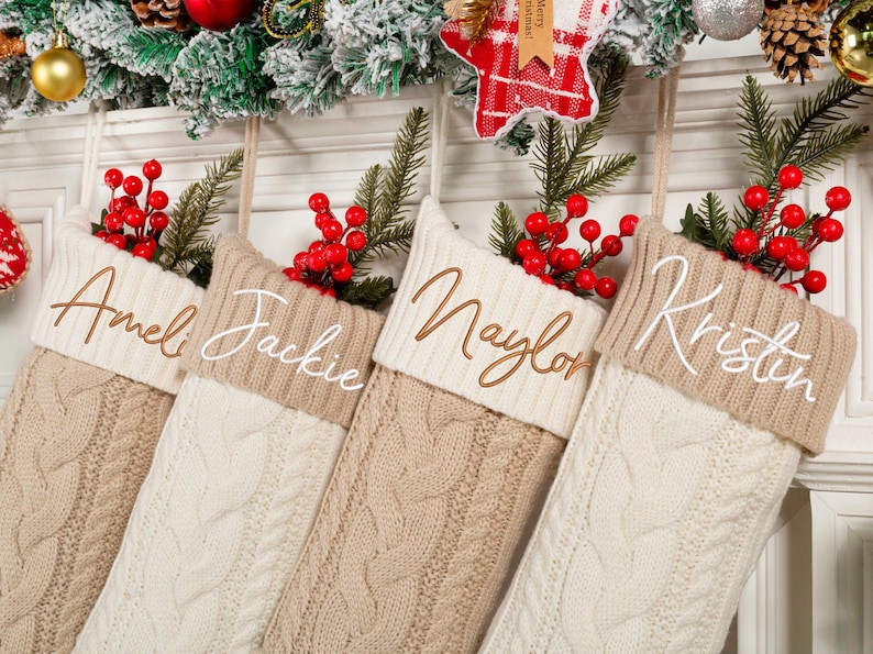 May include: Four cable-knit Christmas stockings in cream and tan, each with a name embroidered on the cuff: Ameli, Jackie, Naylor, and Kristin. The stockings are decorated with red berries and greenery.