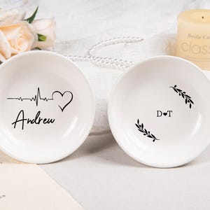 Pode incluir: Dois pratos redondos de cer&acirc;mica branca. O prato esquerdo apresenta uma linha de batimento card&iacute;aco preta, um &iacute;cone de cora&ccedil;&atilde;o e o nome Andrew. O prato direito mostra as iniciais D T separadas por um cora&ccedil;&atilde;o, emolduradas por dois ramos pretos.