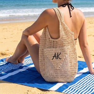 Può includere: Una borsa tote beige all'uncinetto con le iniziali "AK" in nero è mostrata su un telo mare a righe blu e bianche. La borsa ha un ampio design a trama aperta e una tracolla. La scena è ambientata su una spiaggia sabbiosa con l'oceano sullo sfondo.
