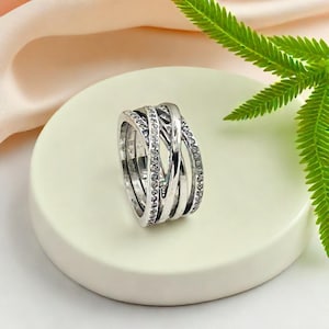 May include: A silver ring with an interwoven design. The ring has multiple bands that cross over each other, with one band decorated with small, clear gemstones. The ring is displayed on a white, circular surface.