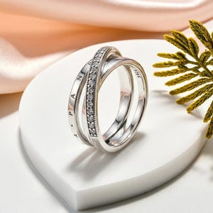May include: A sterling silver crossover ring featuring three bands. One band is pavé-set with clear stones, while another band is engraved with the text PANDORA. The third band is smooth and polished. The interior is stamped with the hallmark S925.