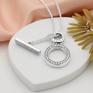 May include: A silver necklace featuring a circular pendant and a bar clasp, both adorned with small, sparkling stones. The necklace is presented on a white surface, set against a soft pink and gold backdrop.