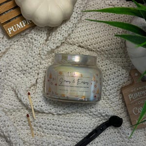 Op de afbeelding: Een heldere glazen kaarsenpot met een parelmoerglans, met het label "S'mores & Embers". De kaars staat op een crèmekleurige gebreide deken, met een witte pompoen en houten accenten. Twee lucifers en een zwarte lonttrimmer zijn ook zichtbaar.