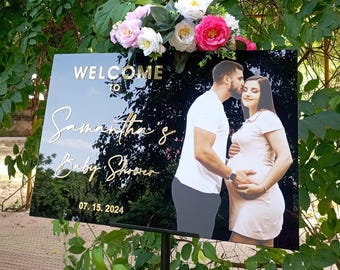 May include: A black acrylic baby shower welcome sign with gold lettering and a photo of a couple. The sign reads "WELCOME to Samantha's Baby Shower" and the date "07.15.2024". The sign is decorated with pink and white flowers.