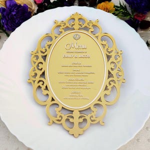May include: An oval gold acrylic menu sign with an ornate, laser-cut filigree frame. The center features the text Menu, Wedding Celebration of Emily & Jacob, and a list of food items including Tomato Basil Soup, Mixed Greens, and Seared Chicken.