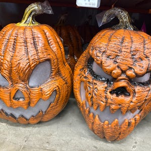 May include: Two orange Halloween pumpkin masks with carved faces. The pumpkins have jagged teeth, glowing eyes, and textured surfaces. The stems are green and wrapped in plastic. Perfect for Halloween.