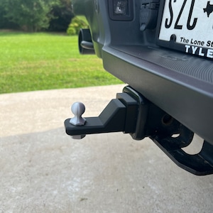 May include: A silver hitch ball attached to a black trailer hitch receiver on a black truck. The truck has a Texas license plate that reads "SZC" with the text "The Lone Star State" and "TYLER" below it.