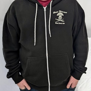 Puede incluir: Una sudadera negra con cremallera y un gráfico blanco en el pecho izquierdo. El diseño muestra una calavera con casco sobre dos espadas cruzadas, con el texto "Deva Warcom" arriba y "Alea Iacta Est" debajo del emblema. La sudadera tiene una cremallera blanca.