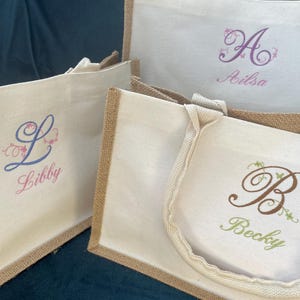 Puede incluir: Tres bolsas de mano personalizadas con tela blanca y ribete de yute. Cada bolsa presenta una inicial y un nombre bordados diferentes: "Ailsa", "Libby" y "Becky". Las bolsas tienen asas resistentes.