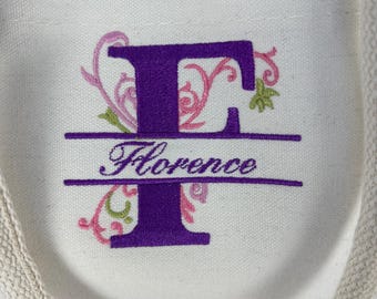 May include: Cream-colored tote bag with a burlap bottom and rope handles. The bag features a large purple letter "F" with floral embroidery in pink and green, and the name "Florence" embroidered below.