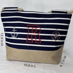 Puede incluir: Bolso tote de temática náutica con rayas azul marino y blancas, bordado con una letra "R" roja, un ancla y una rosa de los vientos. Tiene asas de cuerda y una base beige. Dimensiones: 49,8 cm de largo, 32,8 cm de alto y 15,5 cm de ancho.