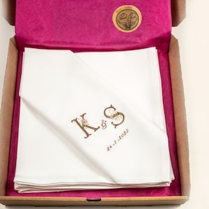 Könnte beinhalten: Gefaltetes weißes Taschentuch mit den gestickten Initialen "K & S" und dem Datum "26.5.2023" in einer braunen Pappschachtel. Die Schachtel ist mit rosa Seidenpapier ausgekleidet. Ein goldener Aufkleber ist sichtbar.