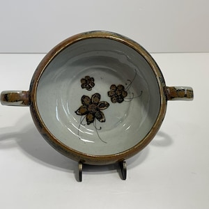 May include: A ceramic bowl with two handles and a floral design. The bowl is off-white inside with brown flowers and black stems. The rim and handles are brown. The bowl is on a metal stand.