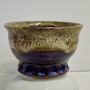 May include: A small, brown and white ceramic bowl with a purple glaze on the bottom. The bowl has a scalloped edge and a slightly flared rim.
