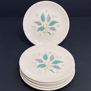 Peut inclure: Ensemble de huit assiettes à salade en céramique blanche avec un motif de feuilles vertes et grises. Les assiettes ont un fond blanc avec un motif de feuilles vertes et grises au centre. Les feuilles sont disposées en forme de bouquet. Les assiettes sont empilées les unes sur les autres.