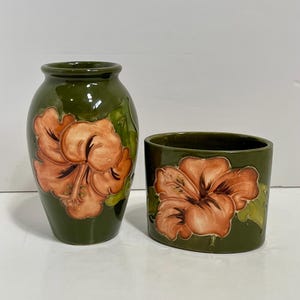 May include: A matching ceramic vase and container set. Both are finished in a dark green glaze and decorated with large, orange hibiscus flower motifs. The vase is tall and narrow, and the container is short and wide.