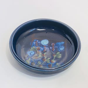 May include: A blue ceramic bowl with a floral design in shades of blue, green, and brown. The bowl has a slightly raised rim.
