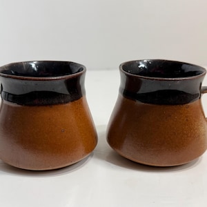 May include: Two ceramic mugs with a wide, rounded base and a tapered top. The lower portion of each mug is a warm, earthy brown, while the upper rim and interior feature a contrasting dark, glossy brown glaze. Each mug has a small side handle.