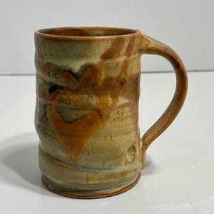 May include: Handmade ceramic mug with a textured surface. The mug features a brown, tan, and green colour palette with a curved handle. The mug is cylindrical and has a rustic, artisanal appearance.