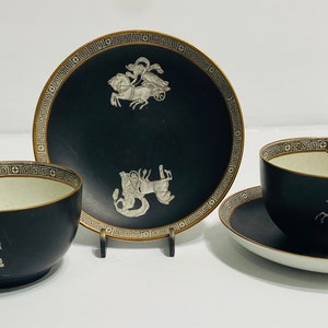 May include: A set of two black teacups and saucers with gold trim and a white Greek-inspired design. The cups and saucers feature a black background with a white design of a chariot pulled by horses.