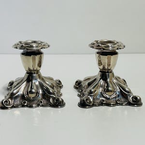 Peut inclure: Deux bougeoirs argentés. Chacun a une base décorative avec un motif floral et un corps cannelé. Le haut de chaque bougeoir a un motif circulaire pour tenir une bougie. Les bougeoirs sont en métal.