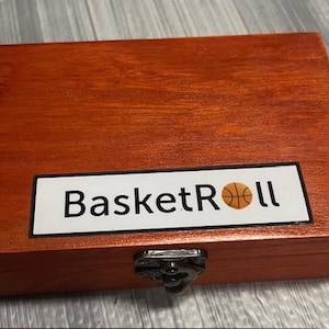 May include: A wooden box with a hinged lid and a latch. The box is stained a reddish-brown colour and has a white label with the text "BasketRoll" and a basketball graphic.