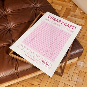 Puede incluir: Una impresi&oacute;n de tarjeta de biblioteca blanca y rosa con las palabras "LIBRARY CARD" en rosa. La impresi&oacute;n est&aacute; en una bandeja de madera sobre un sof&aacute; de cuero marr&oacute;n. La impresi&oacute;n tiene l&iacute;neas para informaci&oacute;n y el texto "Please Return Books on Time".