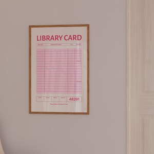 Puede incluir: Una impresi&oacute;n enmarcada de un dise&ntilde;o de tarjeta de biblioteca vintage. La impresi&oacute;n presenta una combinaci&oacute;n de colores rosa y blanco con las palabras "LIBRARY CARD" en la parte superior. La tarjeta tiene l&iacute;neas para informaci&oacute;n y el n&uacute;mero 48291. El texto "Please Return Books on Time" est&aacute; en la parte inferior.
