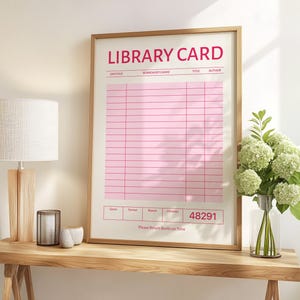 Puede incluir: Una impresi&oacute;n enmarcada de una tarjeta de biblioteca rosa con las palabras "LIBRARY CARD" en rojo en la parte superior. La impresi&oacute;n tiene un marco de madera clara y se exhibe sobre una mesa de madera. Una l&aacute;mpara y un jarr&oacute;n con flores tambi&eacute;n est&aacute;n sobre la mesa.