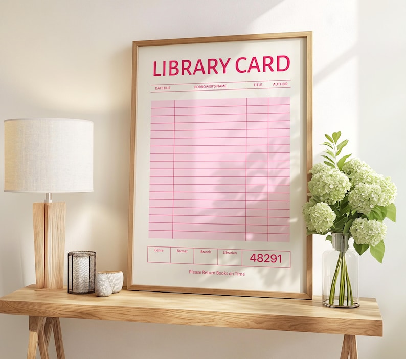 Puede incluir: Una impresi&oacute;n enmarcada de una tarjeta de biblioteca rosa con las palabras "LIBRARY CARD" en rojo en la parte superior. La impresi&oacute;n tiene un marco de madera clara y se exhibe sobre una mesa de madera. Una l&aacute;mpara y un jarr&oacute;n con flores tambi&eacute;n est&aacute;n sobre la mesa.