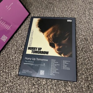 May include: A framed album cover for "Hurry Up Tomorrow" by The Weeknd. The album art features a close-up of a person's face. The album title and artist name are displayed prominently. The tracklist and album details are also visible.