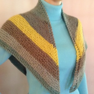 Puede incluir: Un chal triangular de punto con rayas de hilo marrón, gris y amarillo. El chal está drapeado sobre un jersey azul de cuello alto.