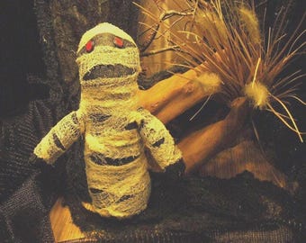 May include: A handmade mummy doll, wrapped in tattered, cream-colored fabric with black stripes. The doll has red eyes and black accents on the head and arms. The Halloween-themed doll is set against a dark background with dried plants.