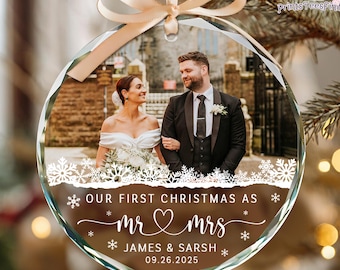 May include: A clear, round Christmas ornament with a photo of a couple. The ornament has a ribbon for hanging and features the text "OUR FIRST CHRISTMAS AS mr & mrs JAMES & SARSH 09.26.2025" with snowflake accents. The background is blurred.