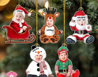 May include: Christmas ornaments featuring babies dressed as Santa, a reindeer, a snowman, and an elf. Each ornament has a baby's name on it. The ornaments are hanging from gold strings against a blurred green background.