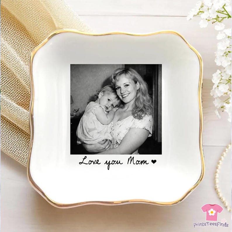 May include: A white ceramic trinket dish with a gold rim and a black and white photo of a mother and child. The dish has the words "Love you Mom" written on it. A perfect gift for Mother's Day.