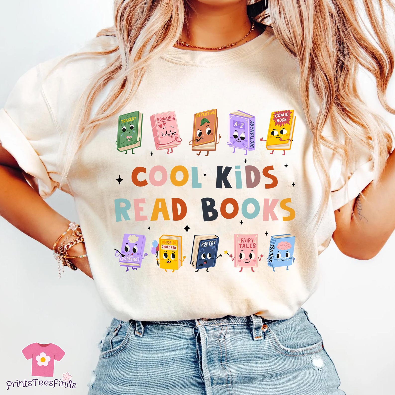 A person wearing a cream-colored graphic t-shirt with the playful slogan "Cool Kids Read Books" printed in a mix of bright, multicolored letters. Above and below the phrase, there are twelve illustrated cartoon books, each with a cute face and tiny limbs, labeled with genres like “Tragedy,” “Romance,” “Detective,” “A–Z Dictionary,” “Comic Book,” “Cooking,” “For Children,” “Poetry,” “Fairy Tales,” and “Science.” The books are arranged in cheerful rows, each expressing different personalities and emotions. The person is wearing light-wash high-waisted jeans and bracelets, with long wavy hair cascading around the shirt. A pink logo in the corner reads “PrintsTeesFinds.”