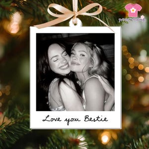 May include: A black and white photo ornament featuring two smiling people embracing. The ornament has a white border and the words "Love you Bestie" are written in cursive at the bottom. A beige ribbon is attached to the top.