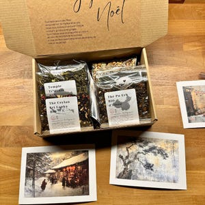Könnte beinhalten: Ein brauner Karton mit den Worten "Joy Noel" auf der Oberseite. Die Schachtel enthält vier kleine Teebeutel mit Etiketten in Französisch. Die Etiketten beinhalten die Namen "Temple Ti-grain", "The Ceylan Sri Lanka BIO", "The Pu Erh BIO d'hiver". Die Schachtel steht auf einer Holzoberfläche mit drei Karten mit Bildern von Winterszenen.