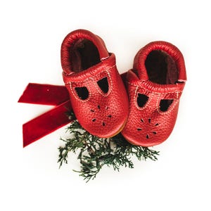 May include: A pair of red leather baby shoes with decorative cutouts and white stitching. The shoes are displayed on a bed of green foliage with a red velvet ribbon, set against a white background. The shoes are a festive red color.